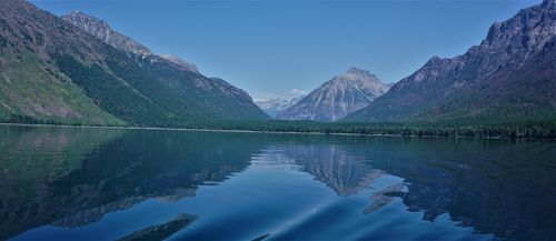 Alt text: A serene lake in Glacier National Park reflects the surrounding forested mountains and jagged peaks under a clear blue sky.