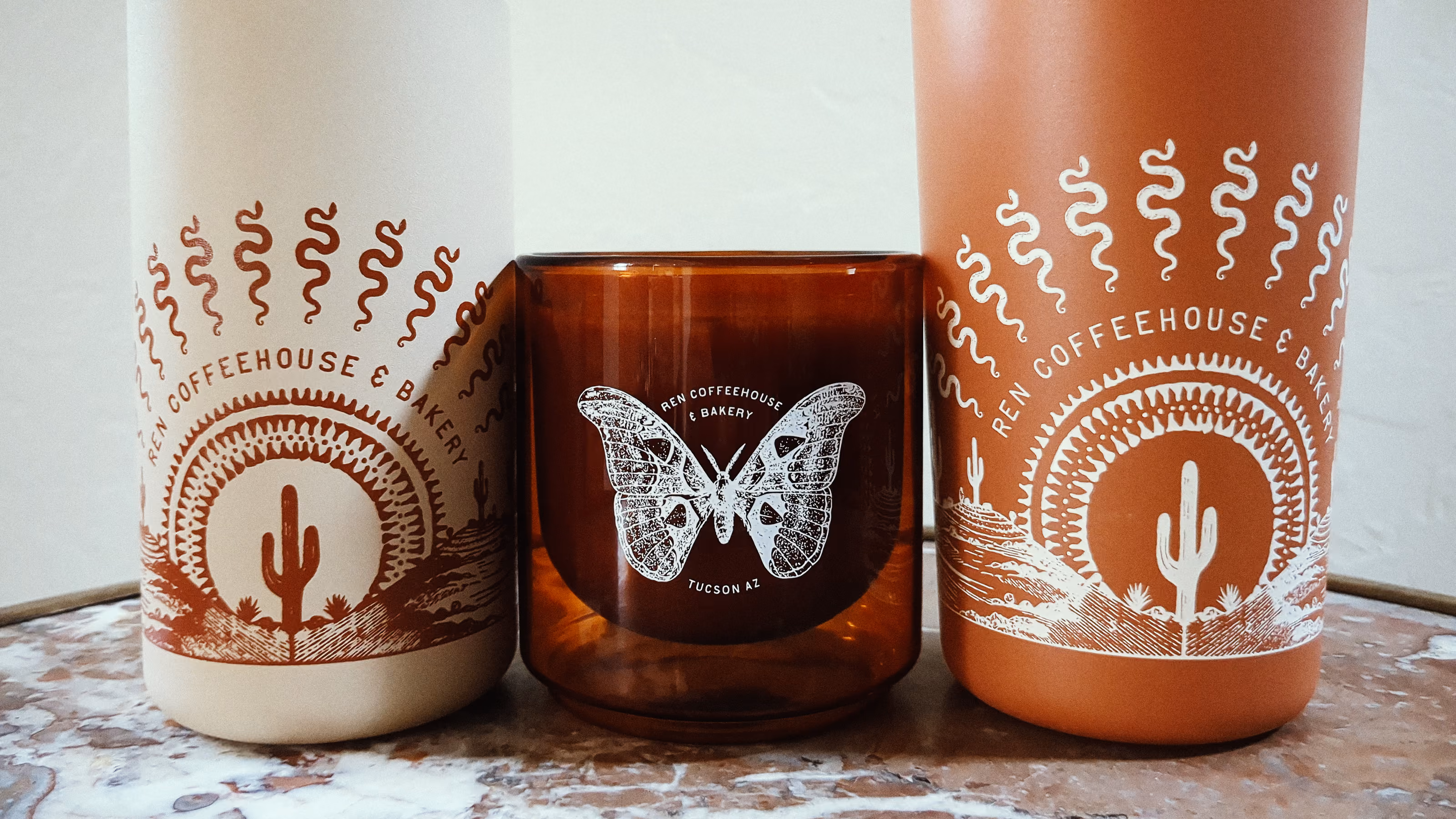 Three ceramic tumblers on a marble surface: two tall ones in cream and terracotta colors with cactus and sun designs labeled 'REN COFFEEHOUSE & BAKERY,' and a shorter amber tumbler in the center with a white butterfly graphic and the same text, placed side by side.