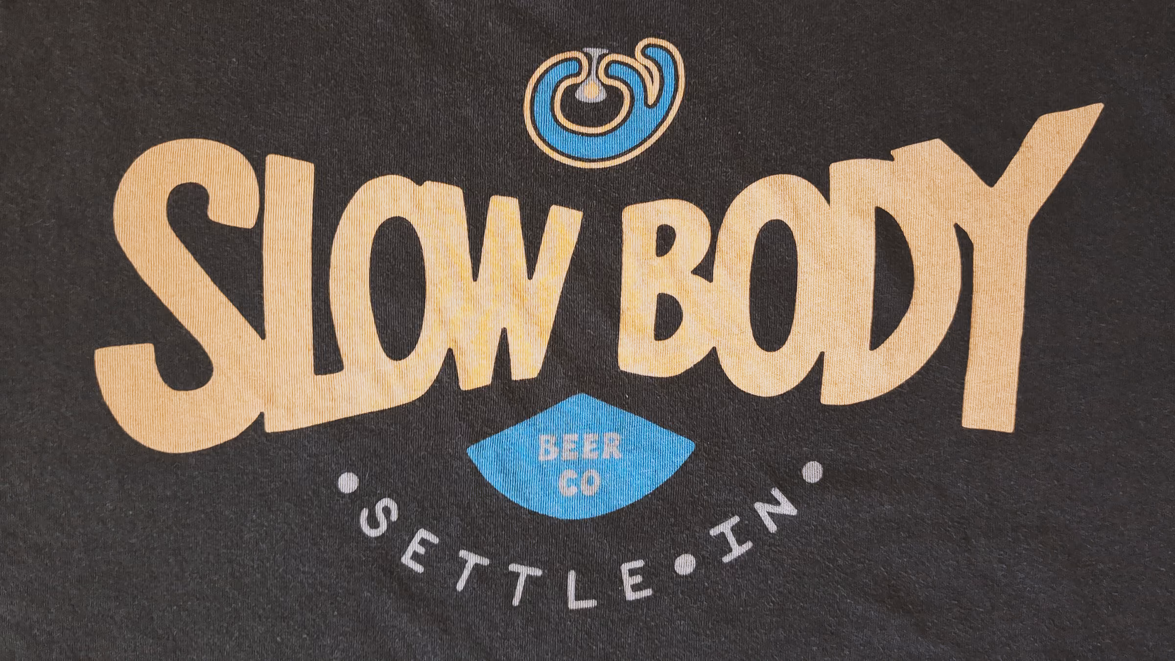 Text design on fabric reading 'SLOW BODY' in large light-colored letters, with a small stylized blue emblem above and a blue diamond below containing the words 'BEER CO' and the phrase 'SETTLE IN' curved underneath.