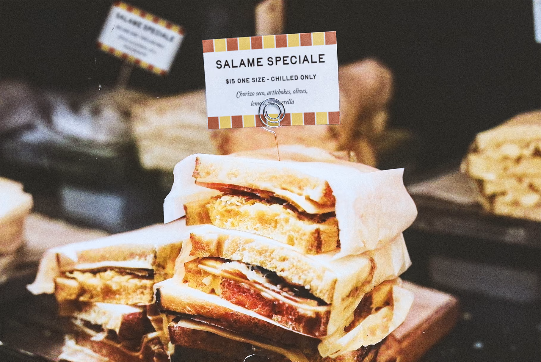 Sandwiches stack one atop another on display in a case with a paper tag in the background