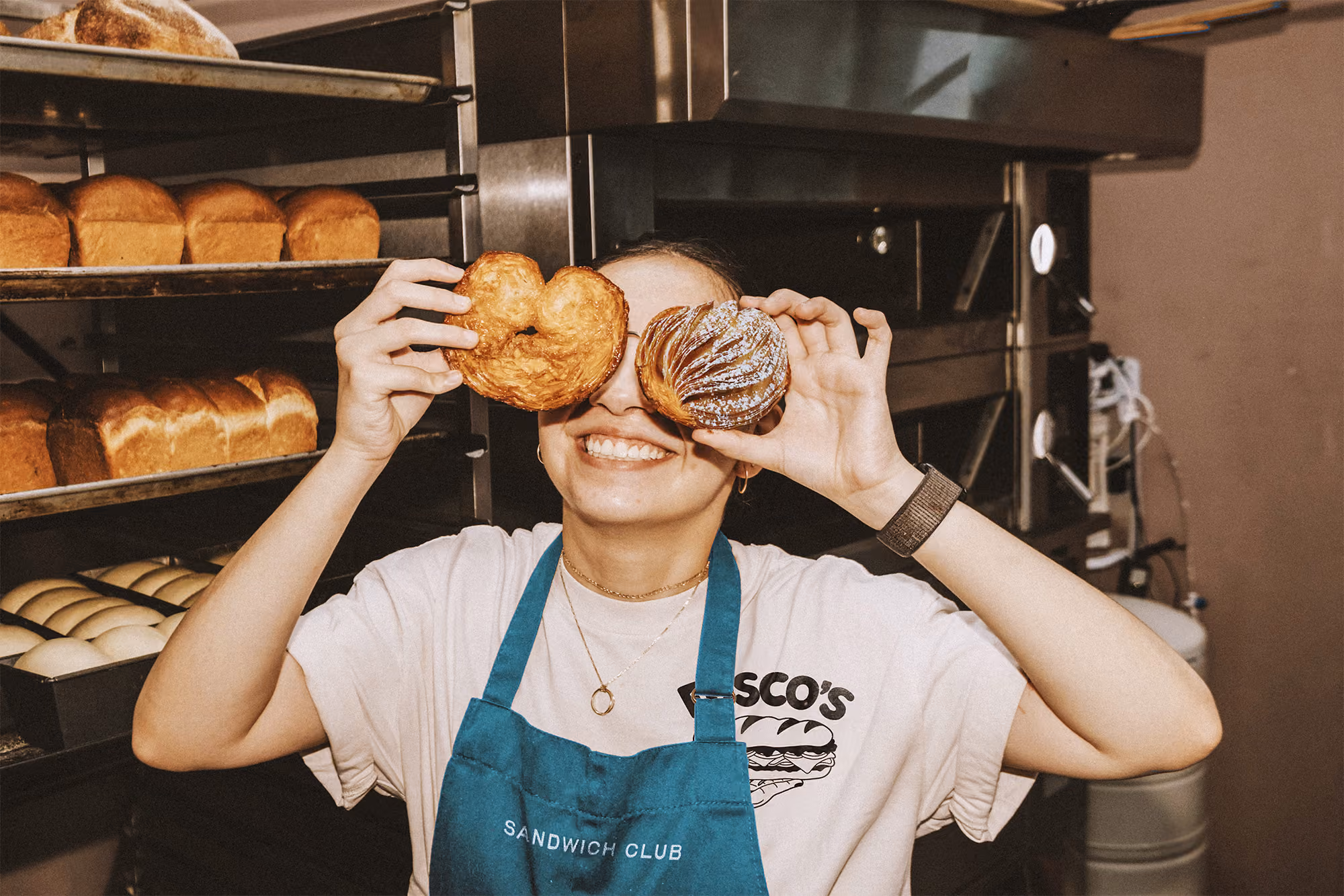 Baker in branded apron holding up 2 pastries in a production space