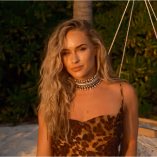 Woman with long blonde wavy hair wearing a leopard-print dress and statement necklace, standing outdoors at sunset with greenery in the background.