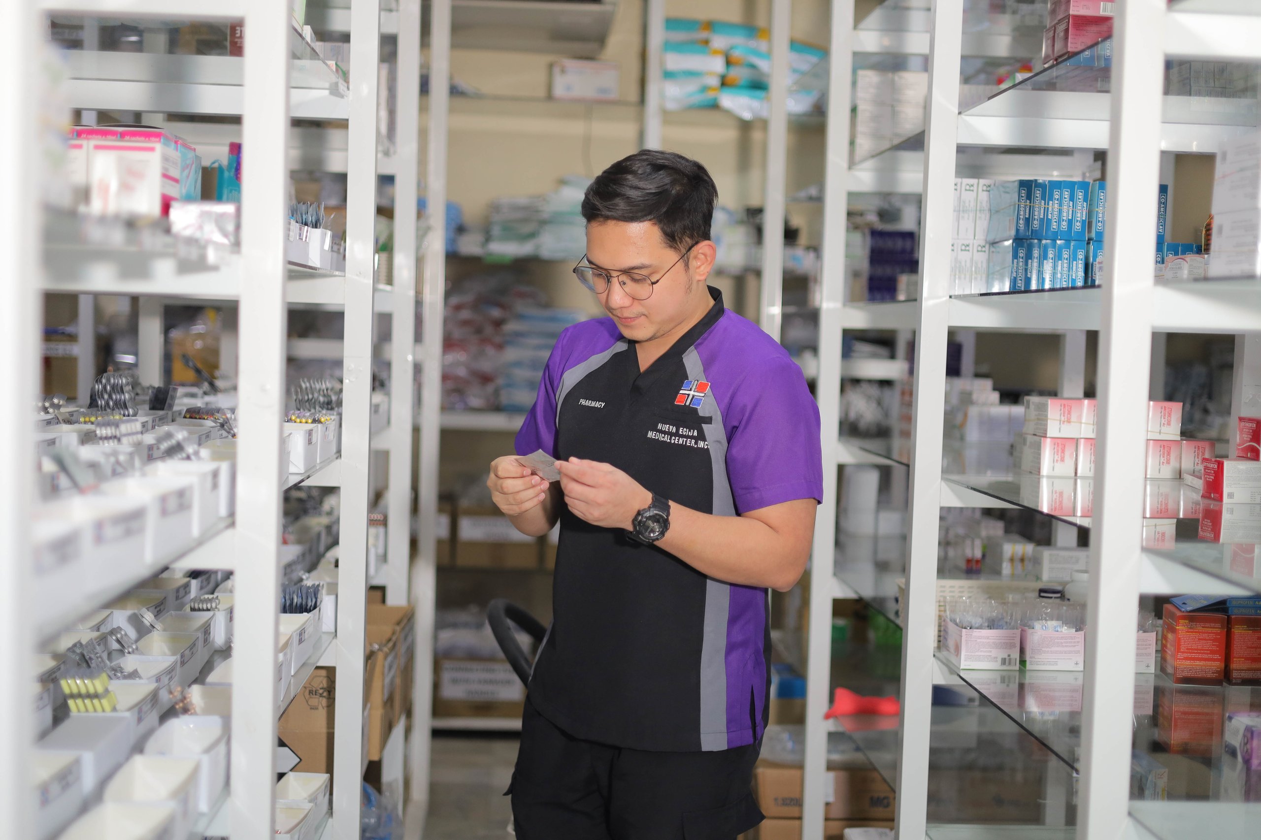 a male pharmacist in a violet and black scrubs  reading a medical prescription in the background are shelves of a pharmacy