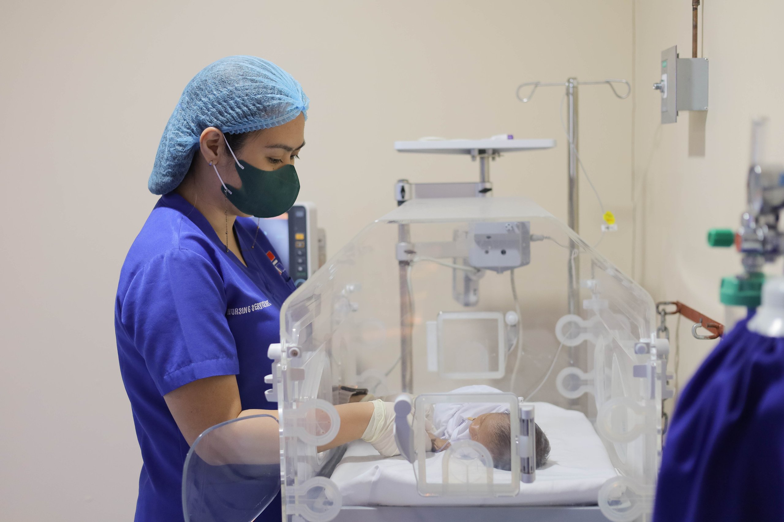 Healthcare worker in blue scrubs and surgical cap caring for a premature infant in a neonatal intensive care unit (NICU) incubator, surrounded by medical monitoring equipment