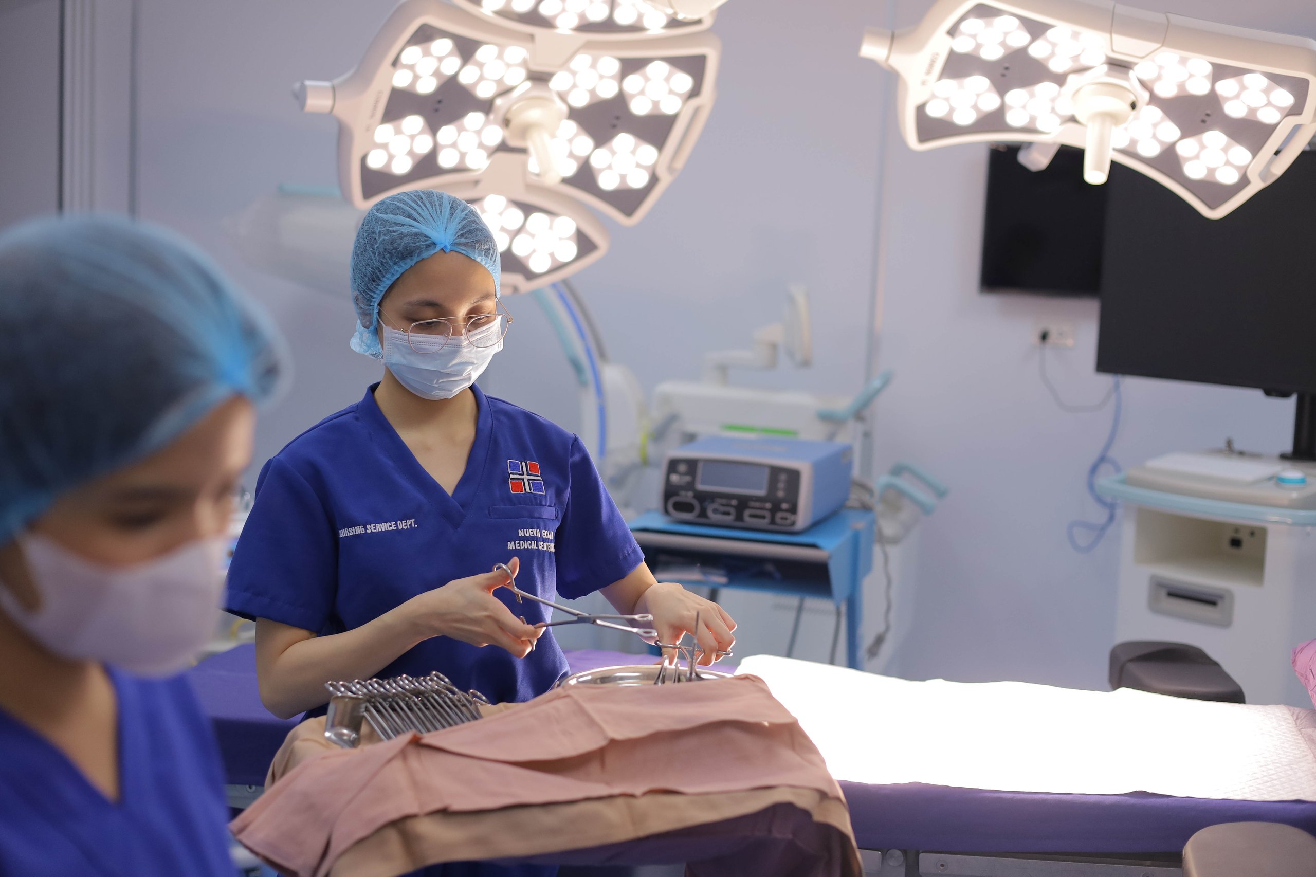 Surgical nurse in blue scrubs, surgical cap, and mask performing a procedure in an operating room under bright surgical lights, with medical equipment and monitors visible in the background