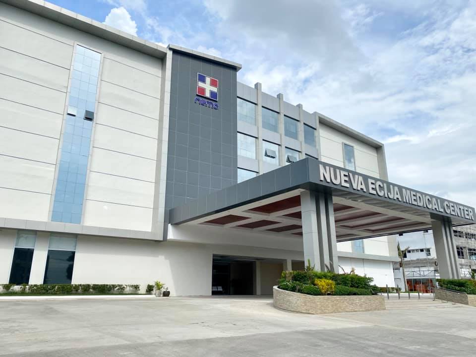 NEMC's Modern three-story hospital building with gray and white exterior, featuring a medical cross logo and 'Nueva Ecija' signage above the covered entrance canopy, set against a partly cloudy sky