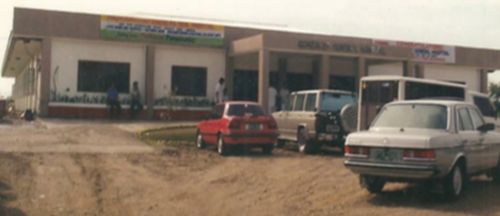 The old Gonzales General Hospital. Vintage photograph showing a single-story commercial or institutional building with cars parked in front, including a red sedan and white SUV on unpaved ground