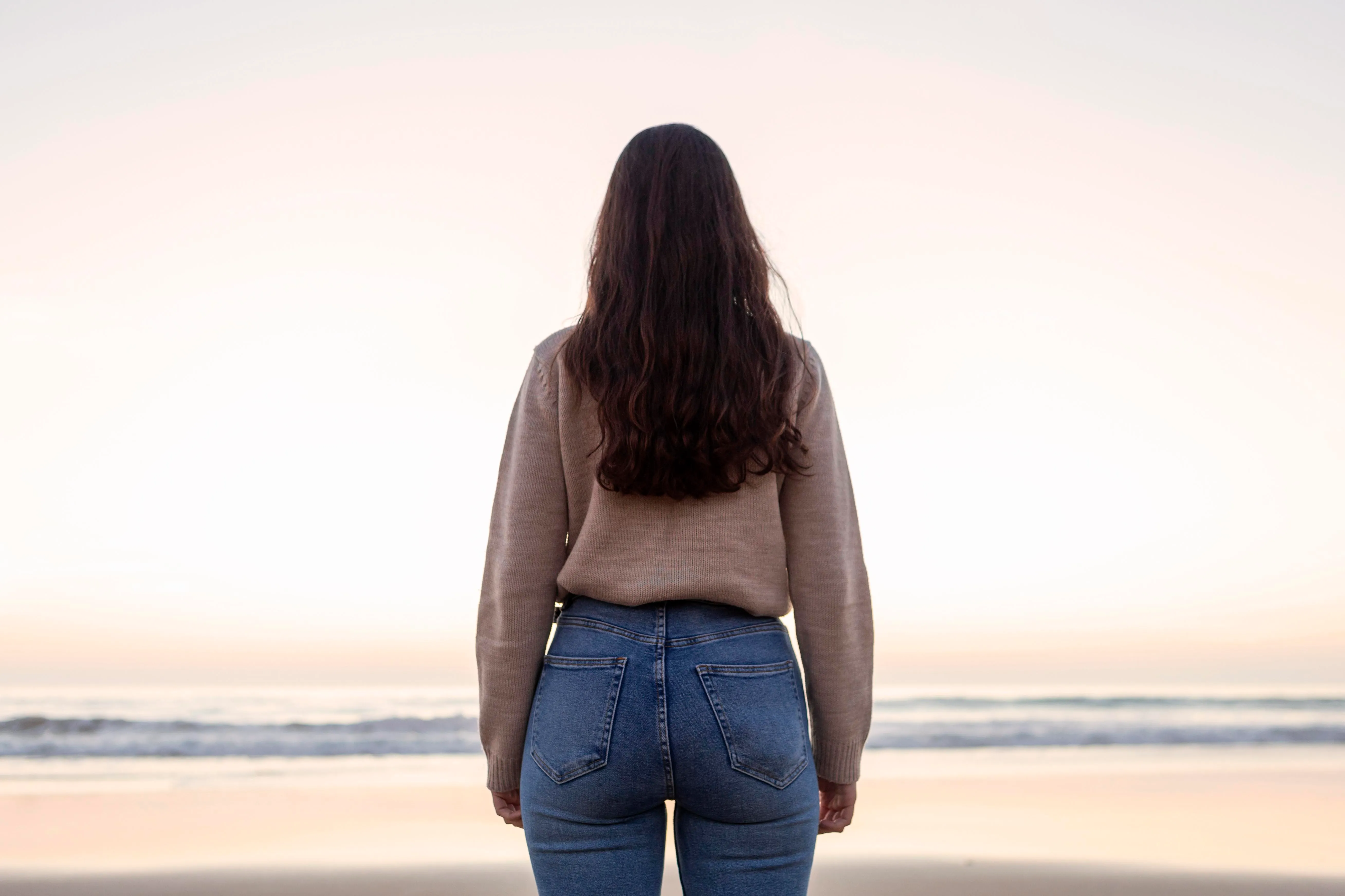 Woman in casual sweater and jeans standing on beach, gazing at ocean waves during sunset, symbolizing calm and reflection in financial decision-making.
