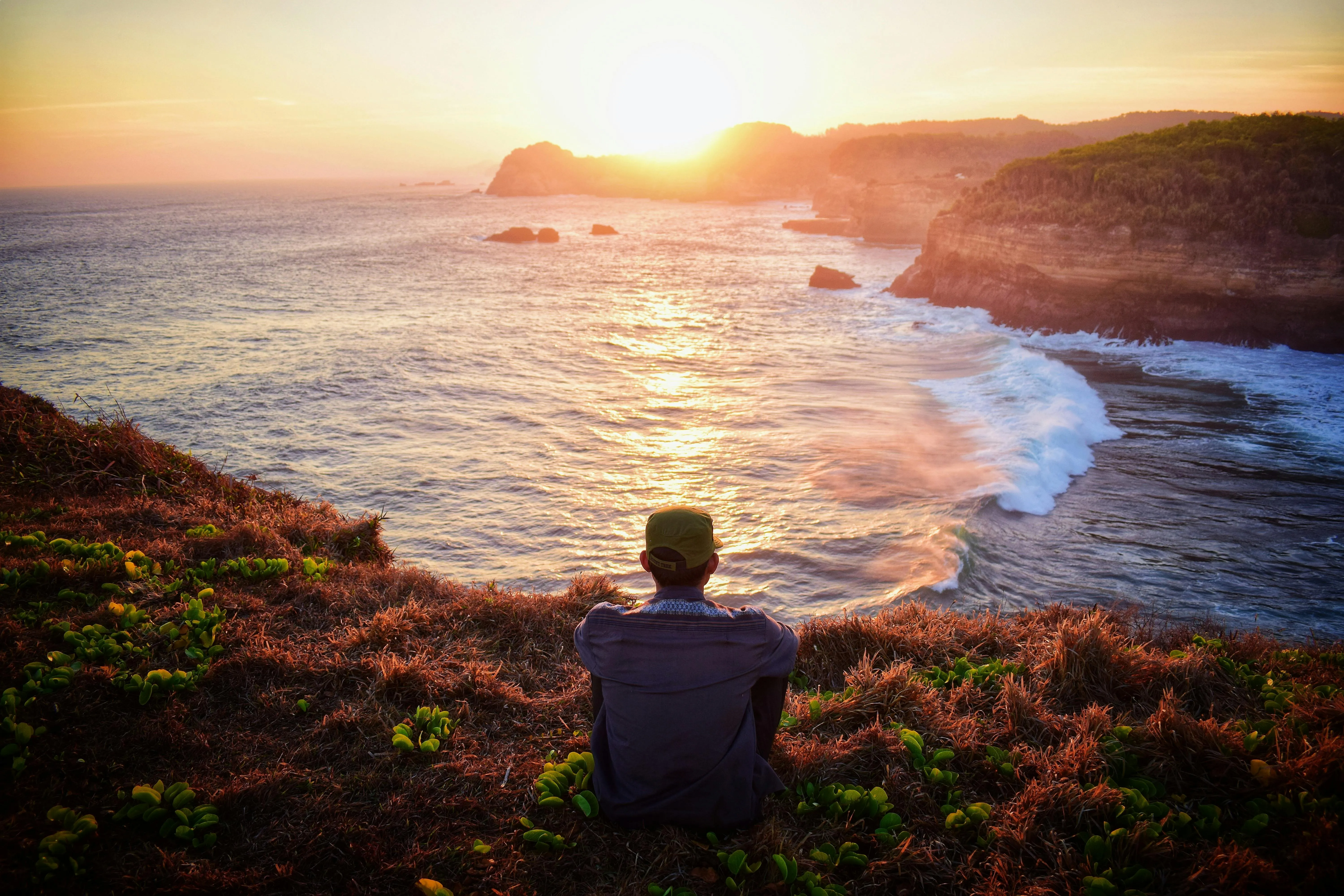 Person sitting on a grassy cliff overlooking the ocean at sunset, reflecting on financial decisions related to Personal Guarantee Insurance.