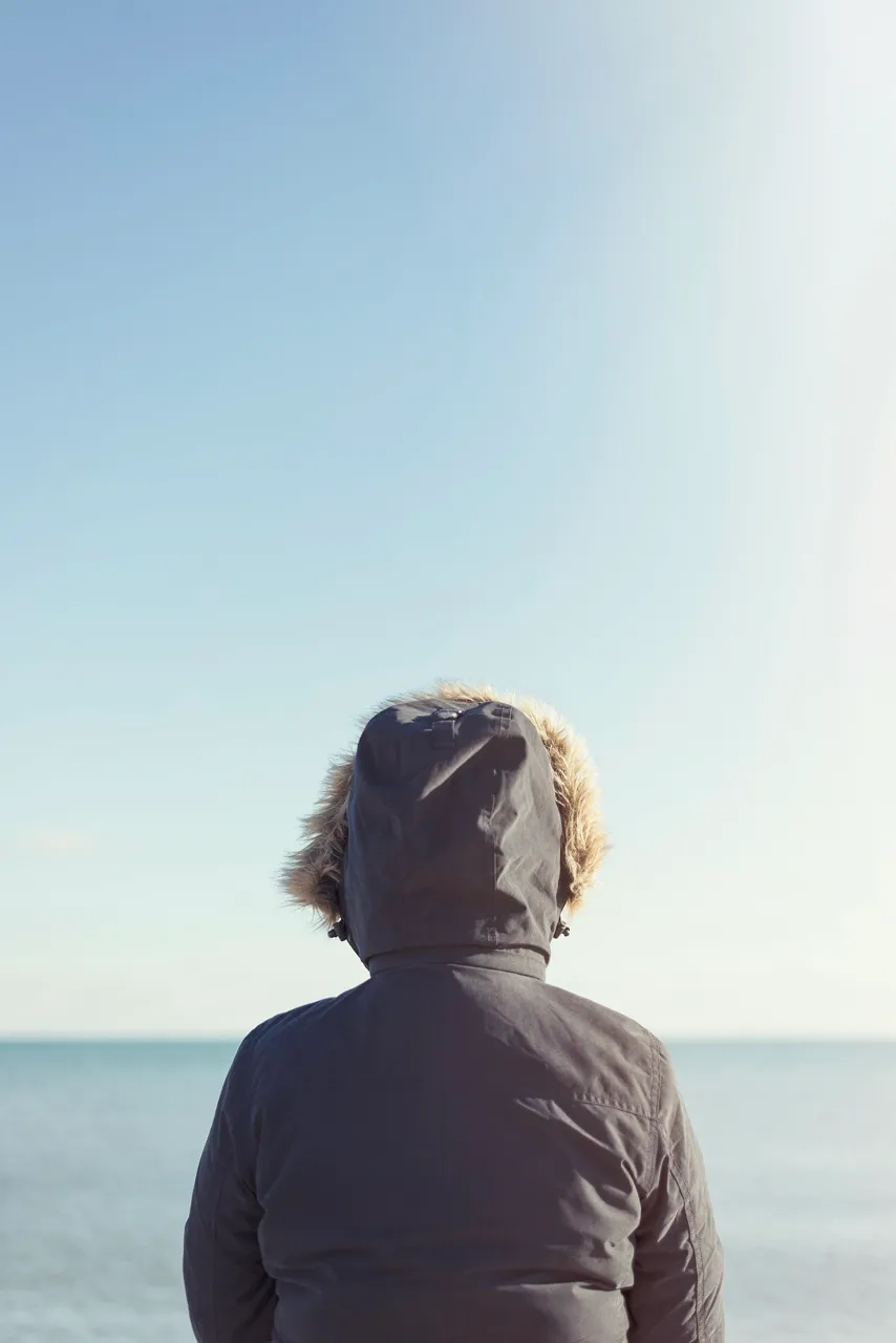 Woman in a coat looking at the sea under a clear sky, reflecting on potential property financing options.