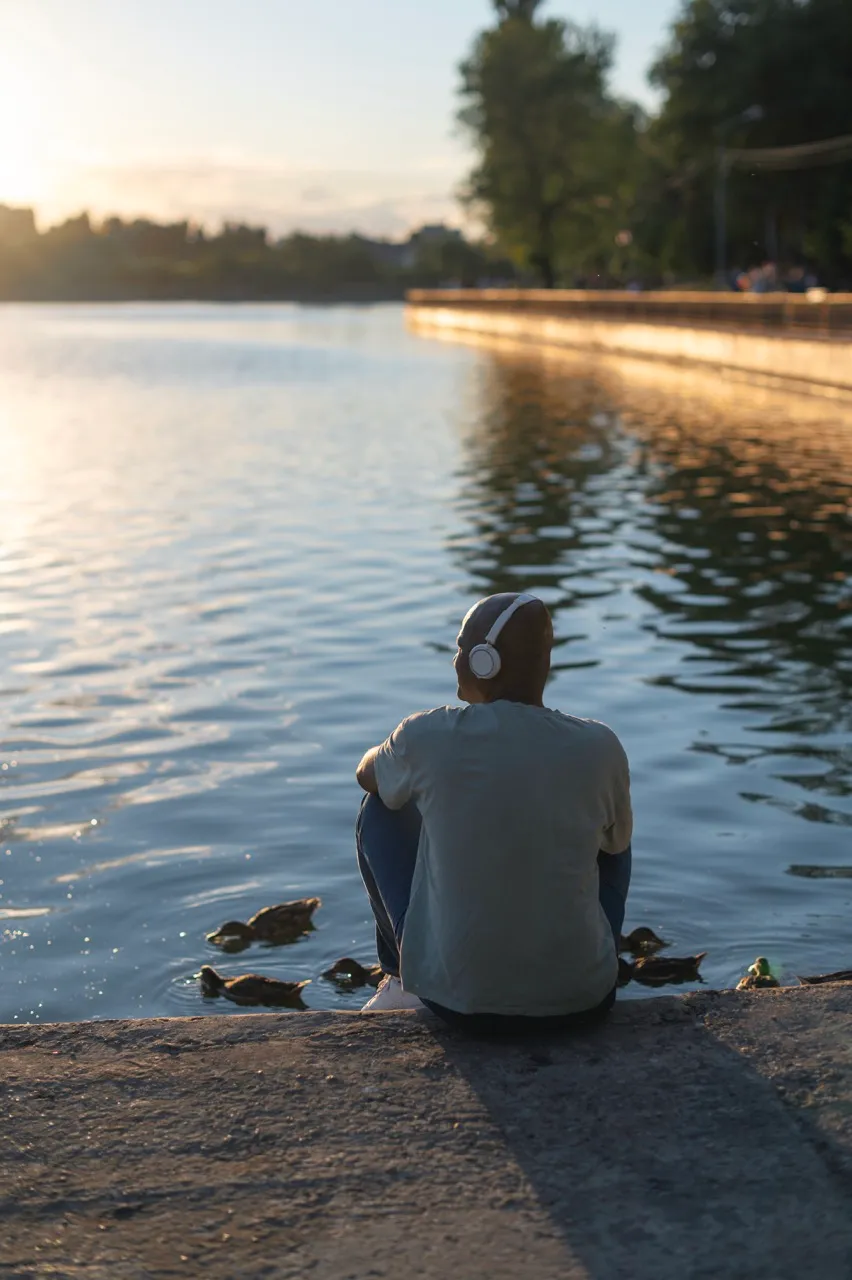 Man sitting by the water wearing headphones, reflecting on asset-based lending opportunities, with ducks swimming nearby in a serene setting.