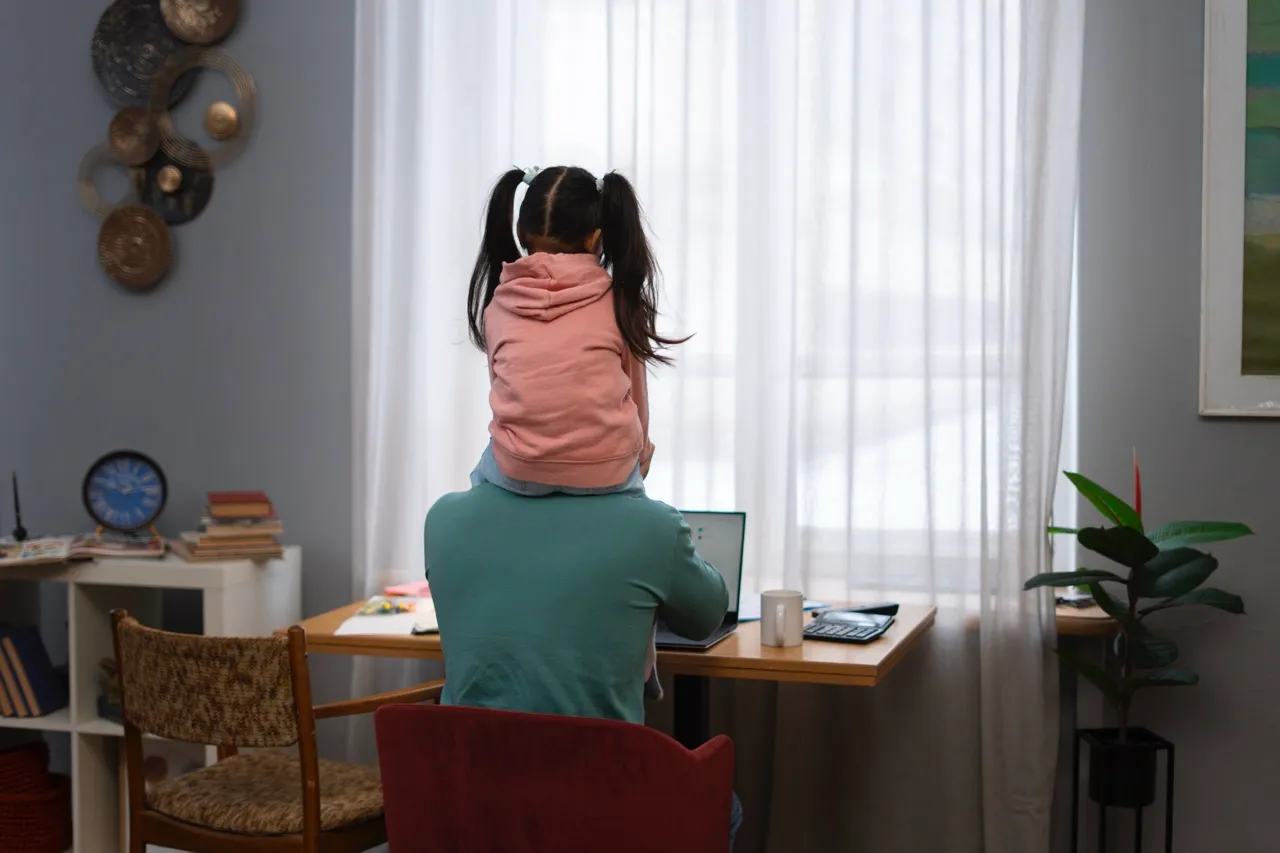 Man working from home on a laptop with a child sitting on his shoulders, surrounded by a cozy indoor workspace featuring a desk, chair, and natural light.