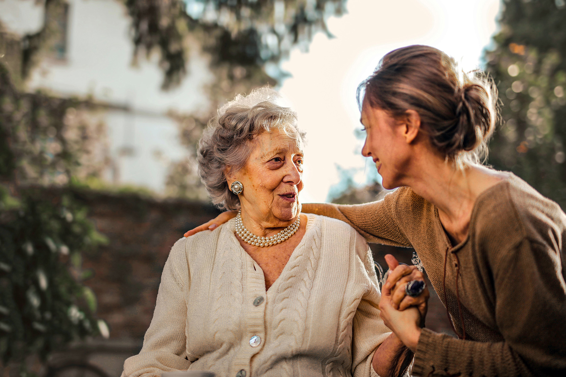 An elderly woman and a younger woman smile at each other while holding hands outdoors, sharing a warm, joyful moment