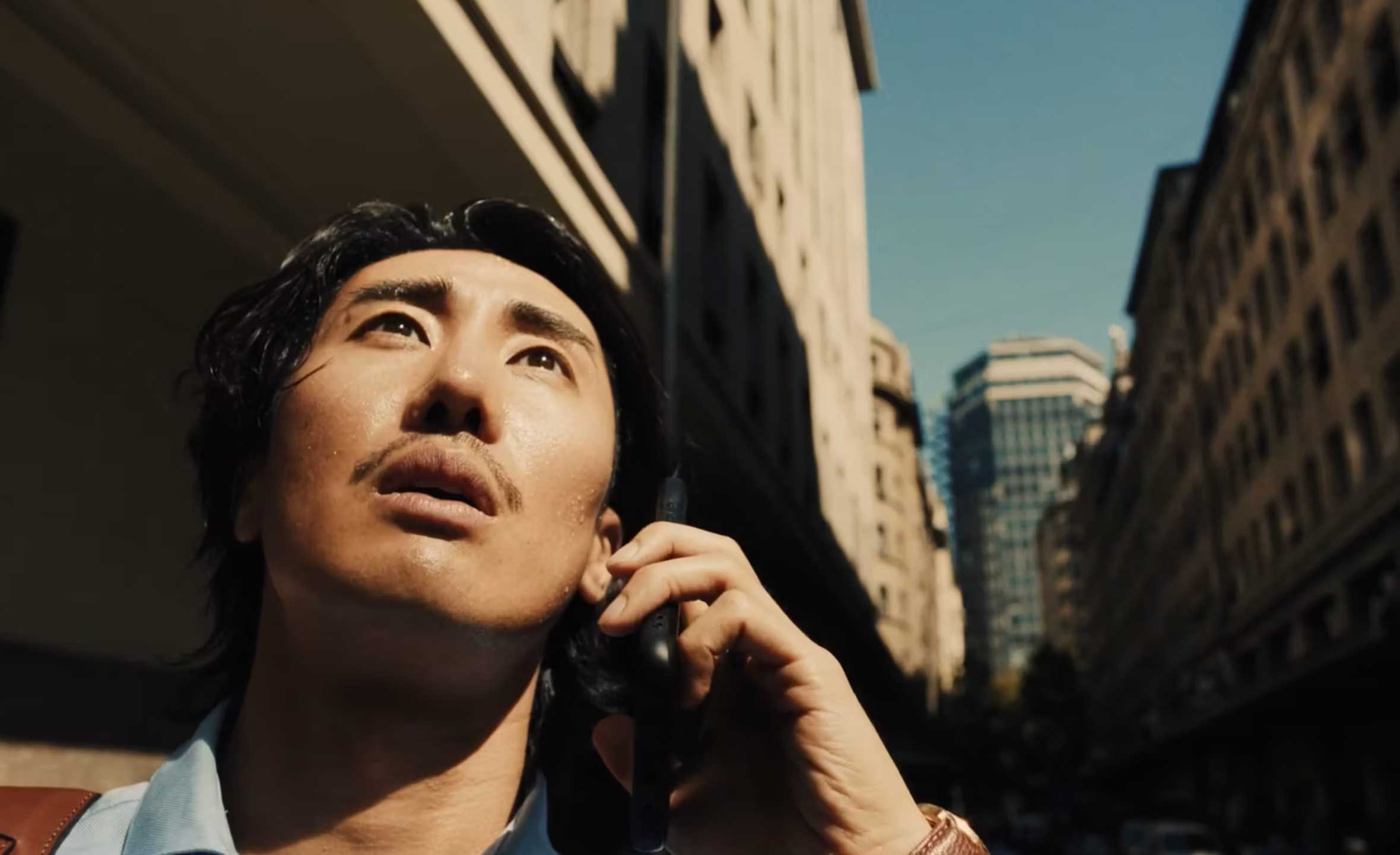 A man holding a phone looks up towards the sky in an urban setting, surrounded by tall buildings and clear blue skies.