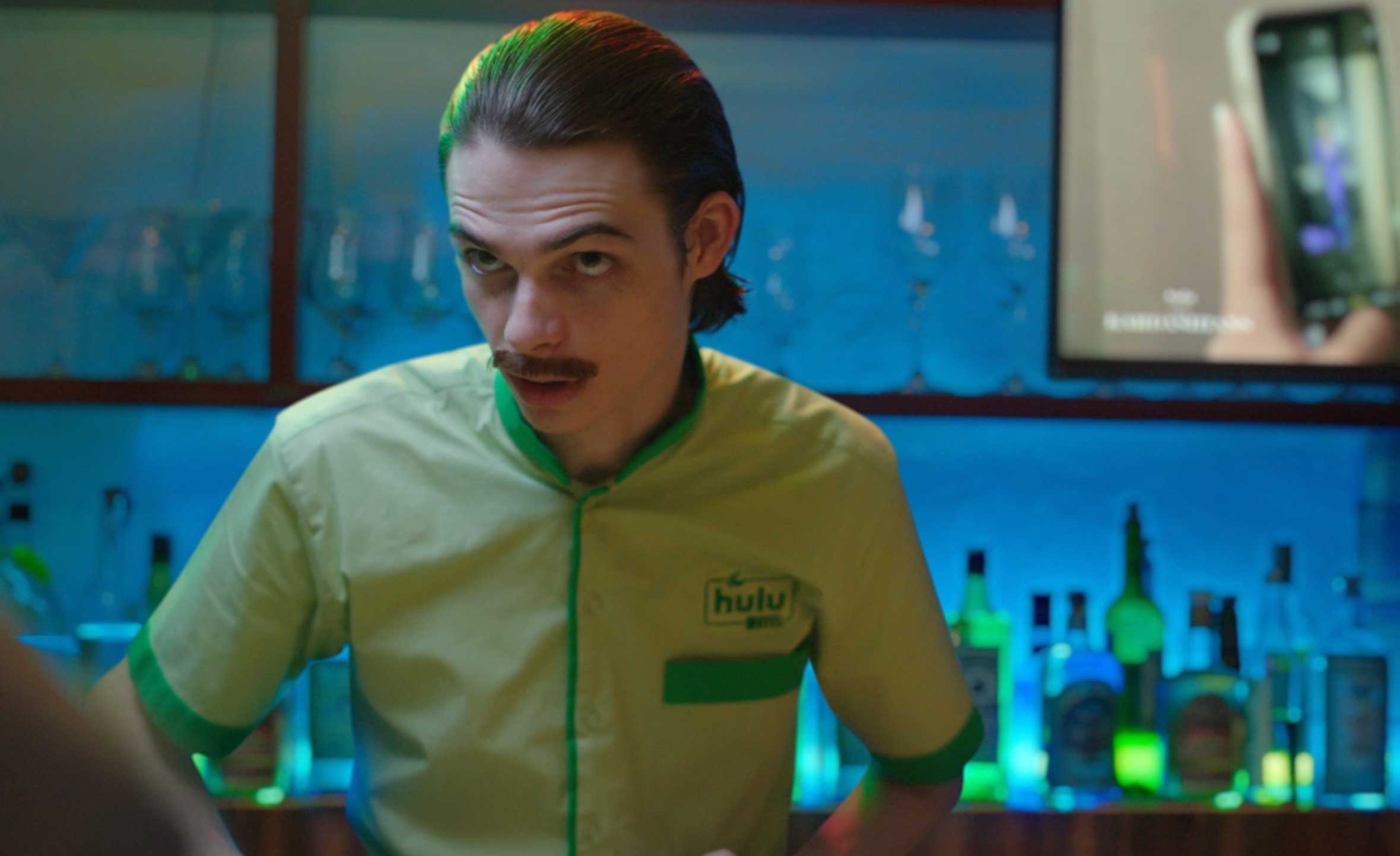 A bartender in a green and yellow uniform stands behind a bar with various bottles, illuminated by colorful lights.
