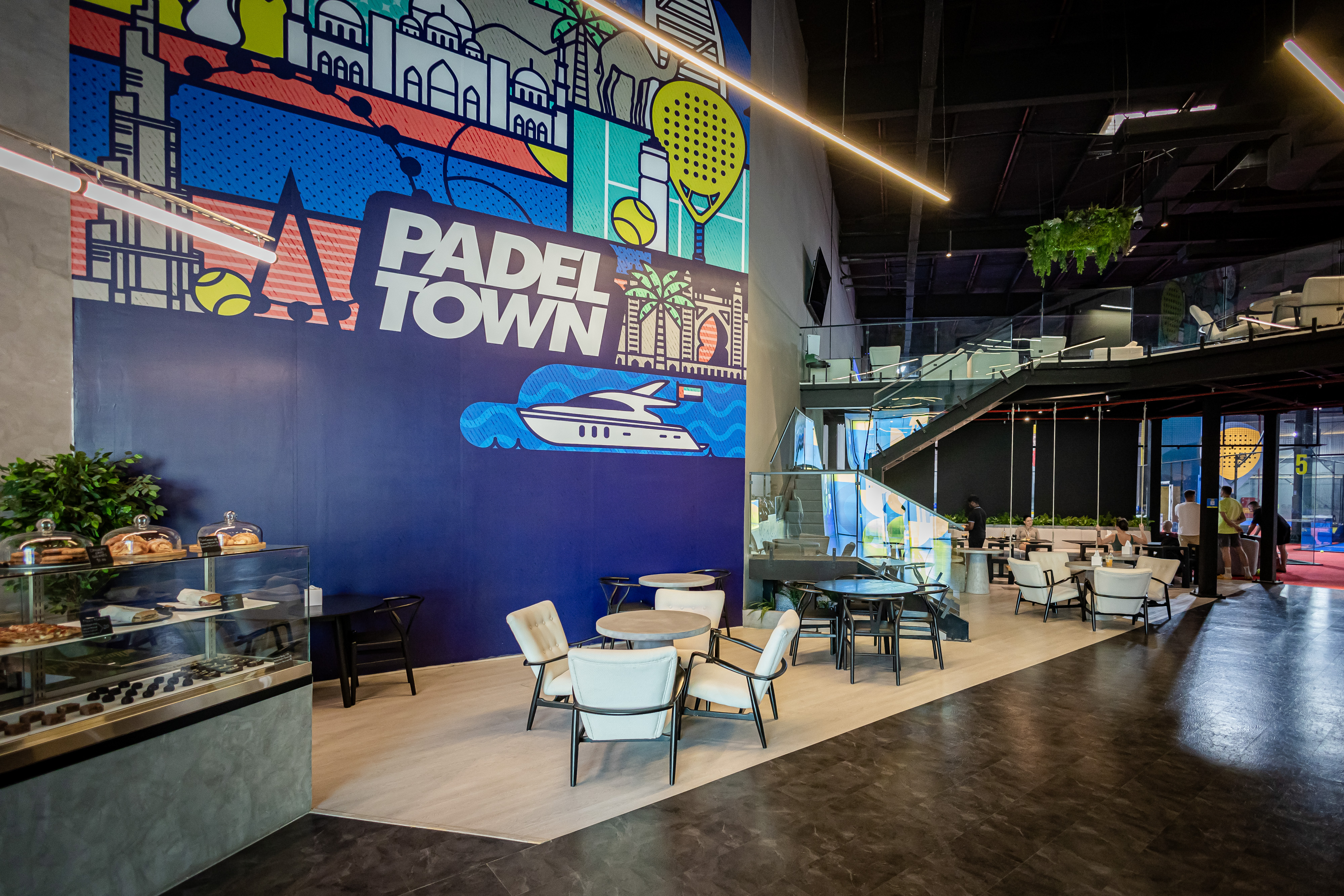 Modern indoor seating area with cushioned chairs and tables under a mezzanine, featuring a large colorful mural at Padel Town.