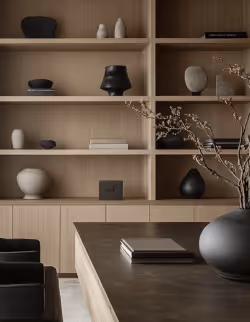 Moderner Raum mit Holzregalen, die minimalistische Keramikobjekte und Bücher zeigen, mit einem Tisch und einem großen schwarzen Vasenarrangement im Vordergrund.