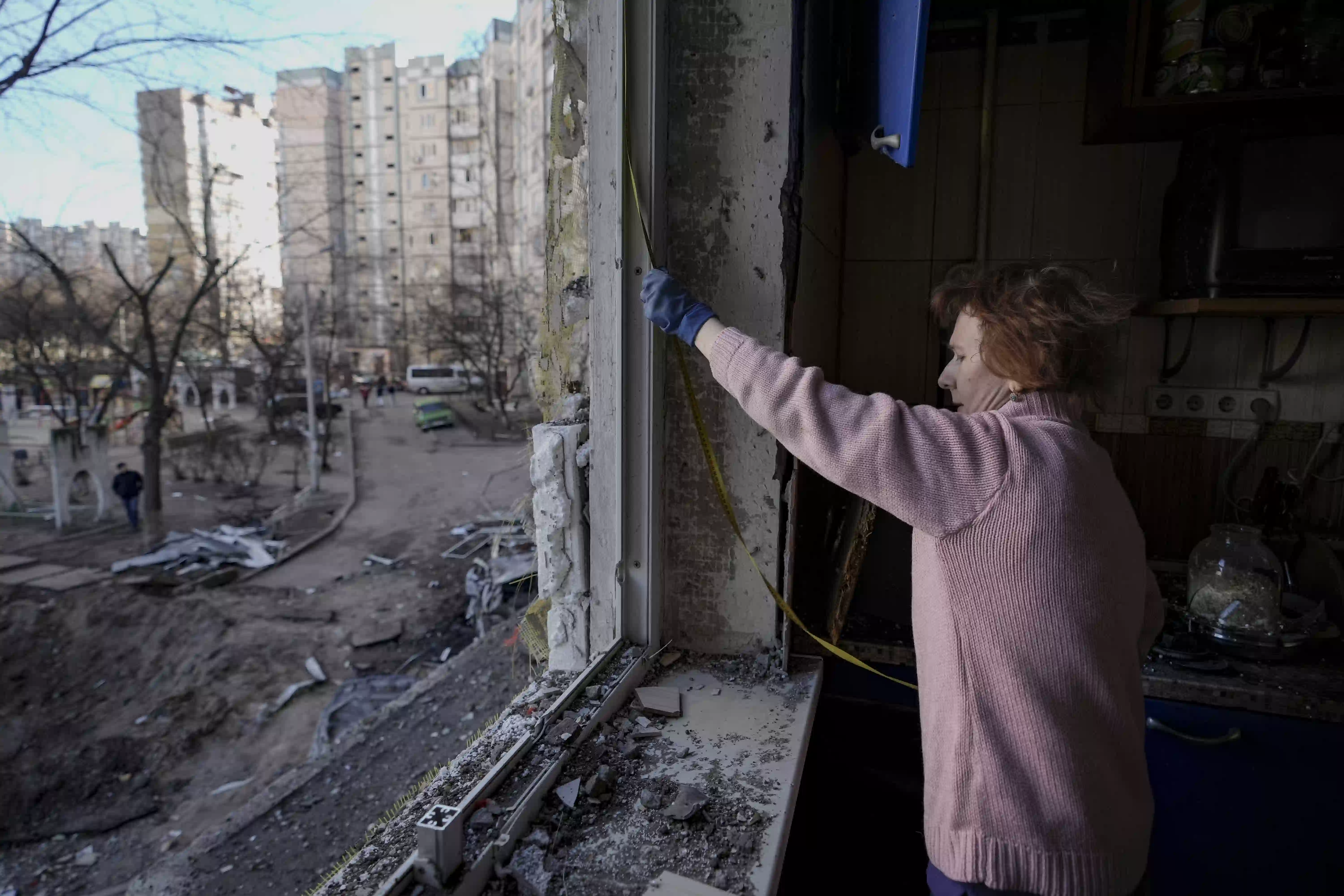 Frau in rosa Pullover misst zerstörtes Fenster in einer beschädigten Wohnung.