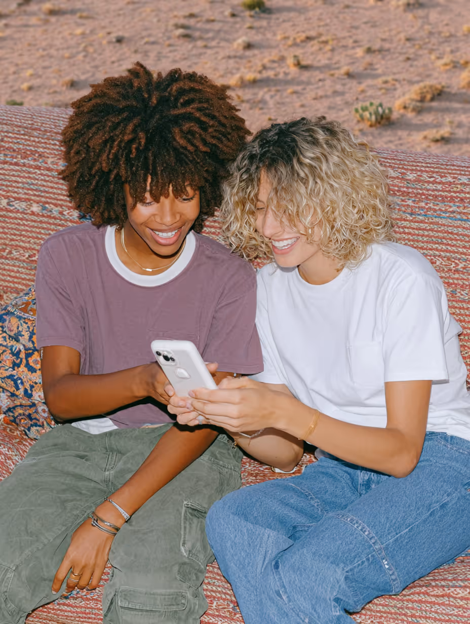 Zwei lachende Personen sitzen zusammen auf einem Teppich und schauen auf ein Smartphone.
