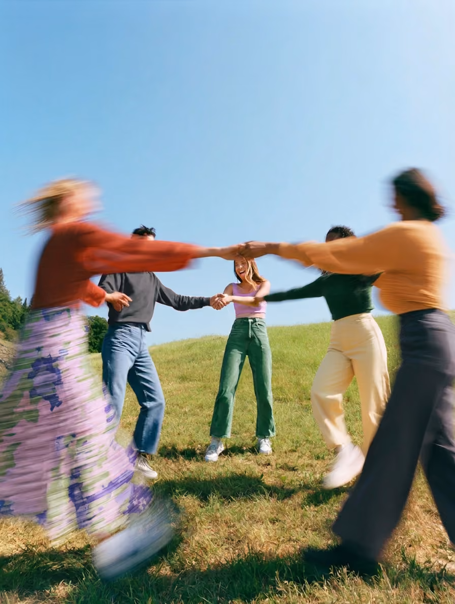 Gruppe lachender Menschen, die sich im Kreis im Freien bei sonnigem Wetter an den Händen halten.
