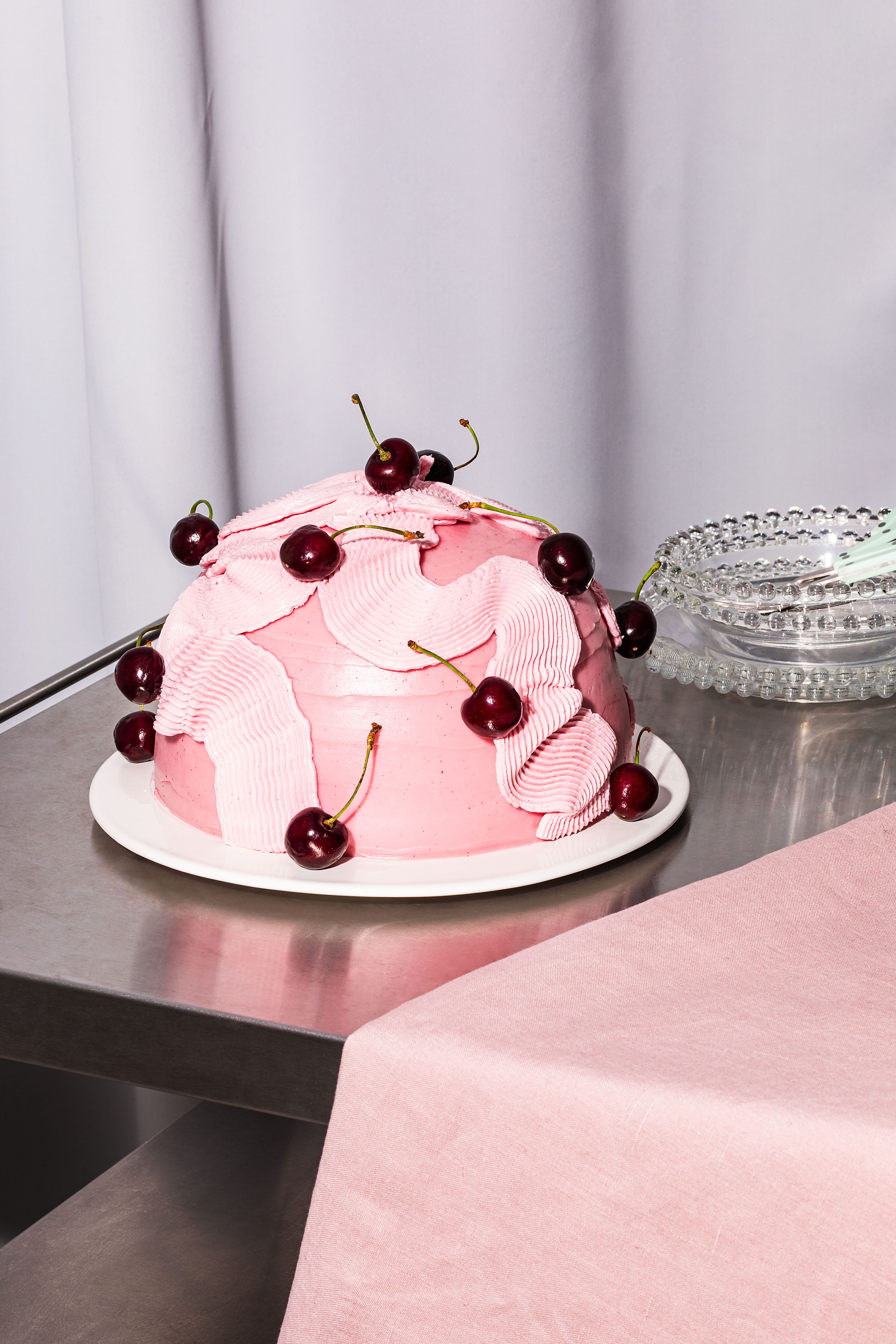 Runde pinke Torte mit dekorativer Glasur und Kirschen auf weißem Teller, silberner Tisch.