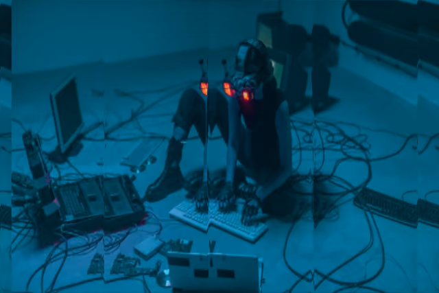 Person mit leuchtenden roten LED-Masken sitzt auf dem Boden umgeben von Kabeln und Computern.