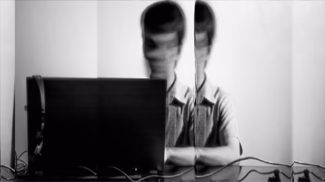 Schwarz-weiße verzerrte Aufnahme einer Person hinter einem Laptop, reflektiert auf einer Glasfläche.