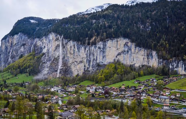 Berglandschaft mit Wasserfall über grünem Tal und Dorf unter bewaldeten Felsen.