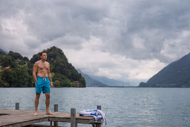 Mann mit Badehose steht auf einem Steg vor einem See und bewölktem Bergpanorama.