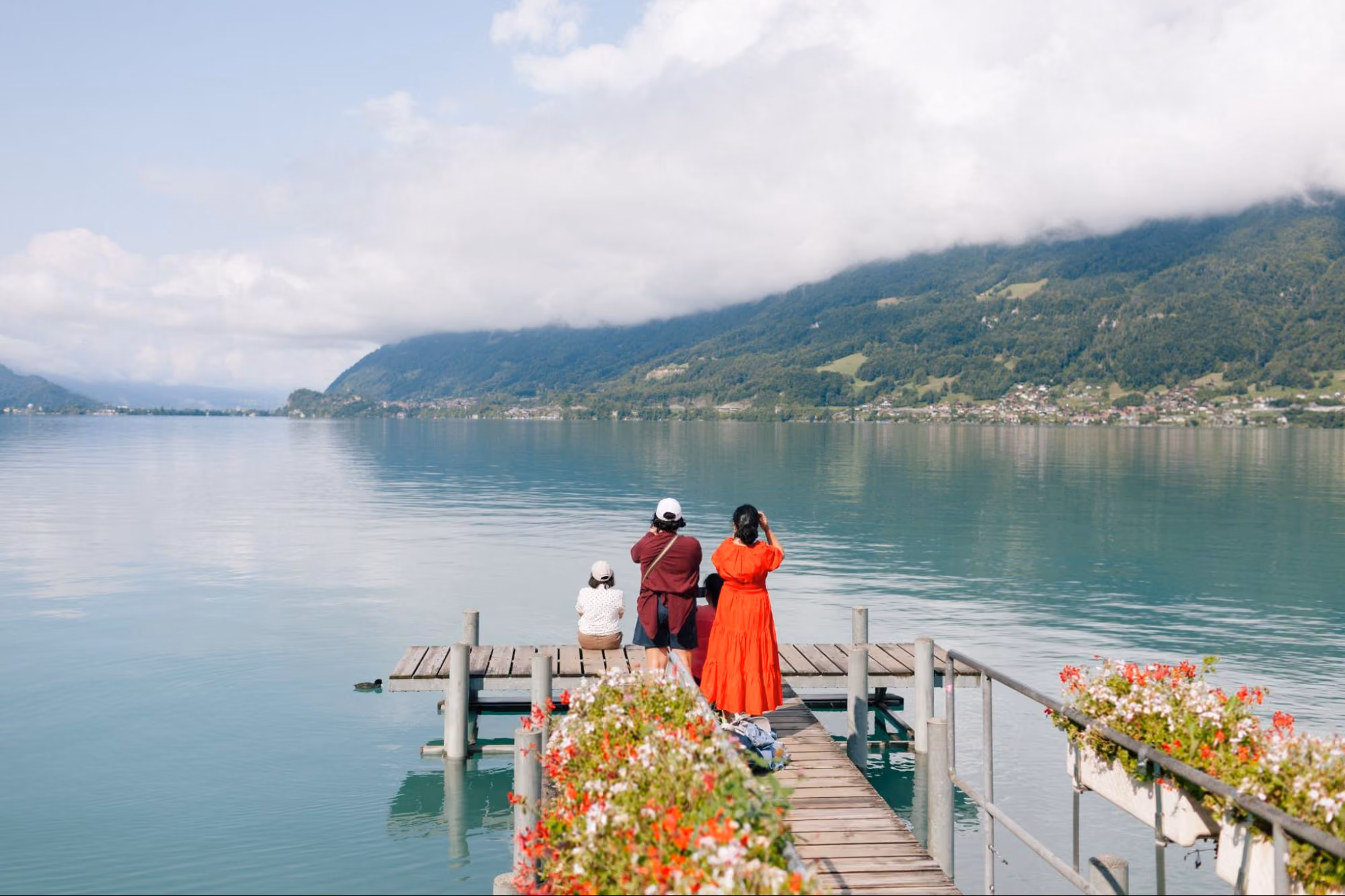Drei Personen auf einem Steg schauen auf einen ruhigen See mit bewaldeten Bergen unter Wolken.