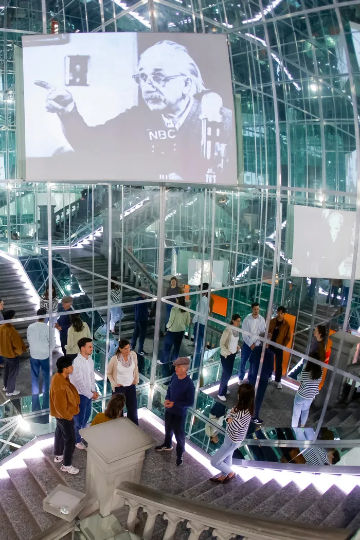 People under reflective mirror walls, a film about Albert Einstein is being projected.