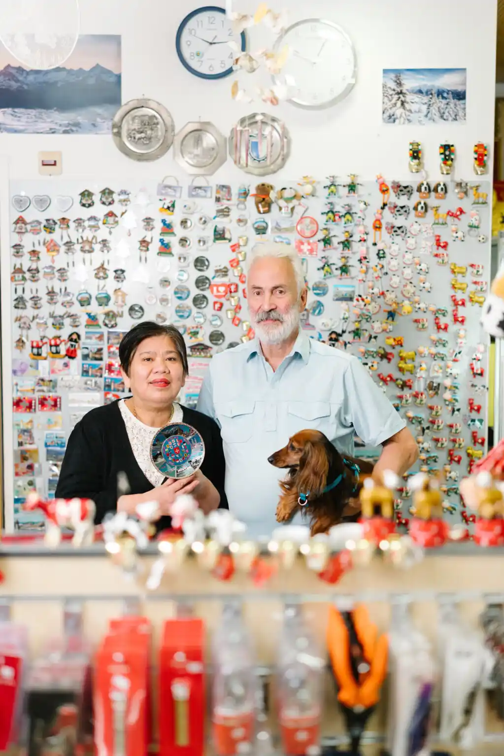 A couple in a souvenir shop with many magnets on the wall and a dog in the man's arms.