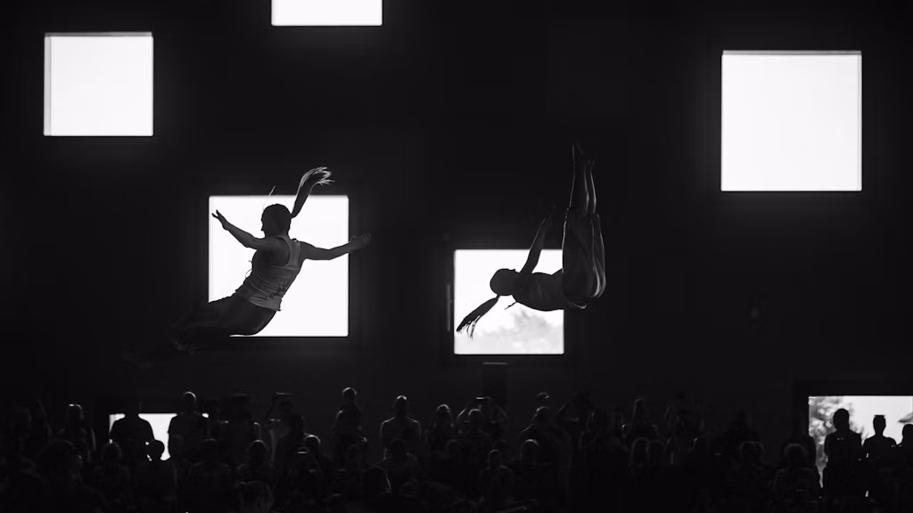 Two gymnasts jumping in a hall with large window blocks, audience in the background.