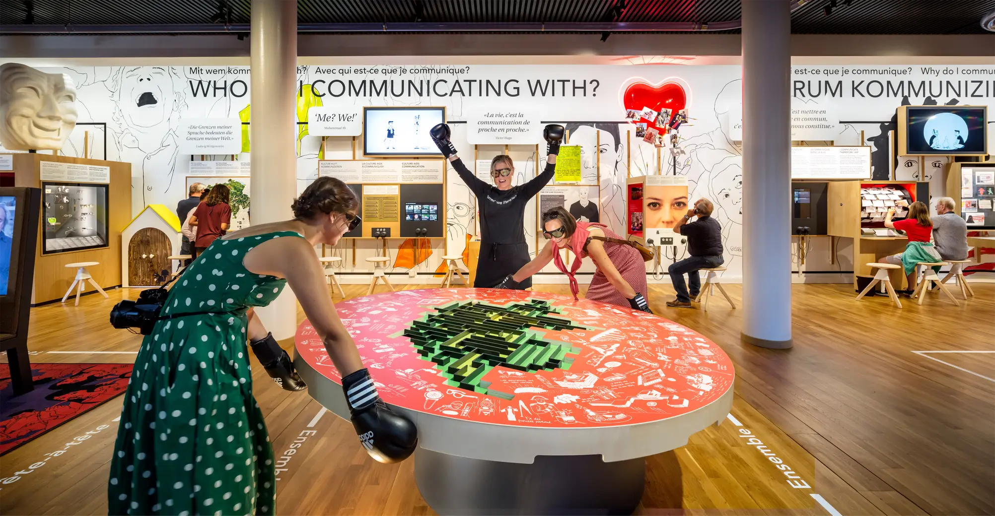 Three people wearing boxing gloves playing at a large red table with a green 3D maze.