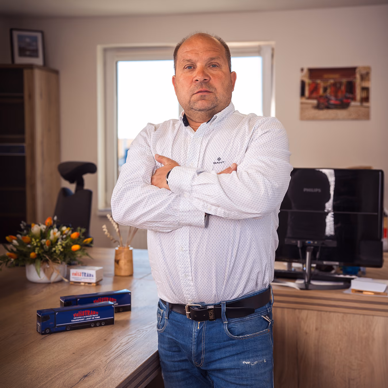 Muž se sklopenýma rukama v kanceláři s modelem nákladního auta na stole a počítačem v pozadí.