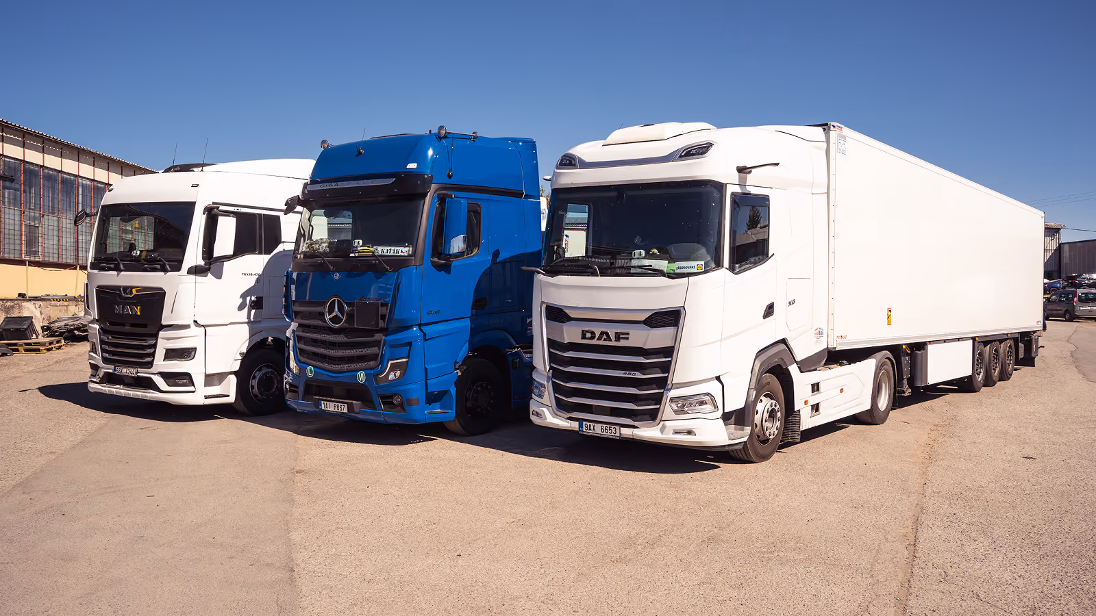Tři nákladní auta značek MAN, Mercedes a DAF zaparkovaná vedle sebe na slunečném dvorku.