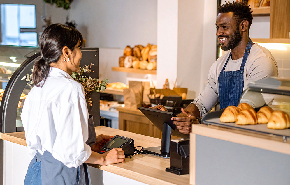 Butcher wearing protective glove and apron holding a large raw steak and knife while a customer pays with cash at a meat counter.
