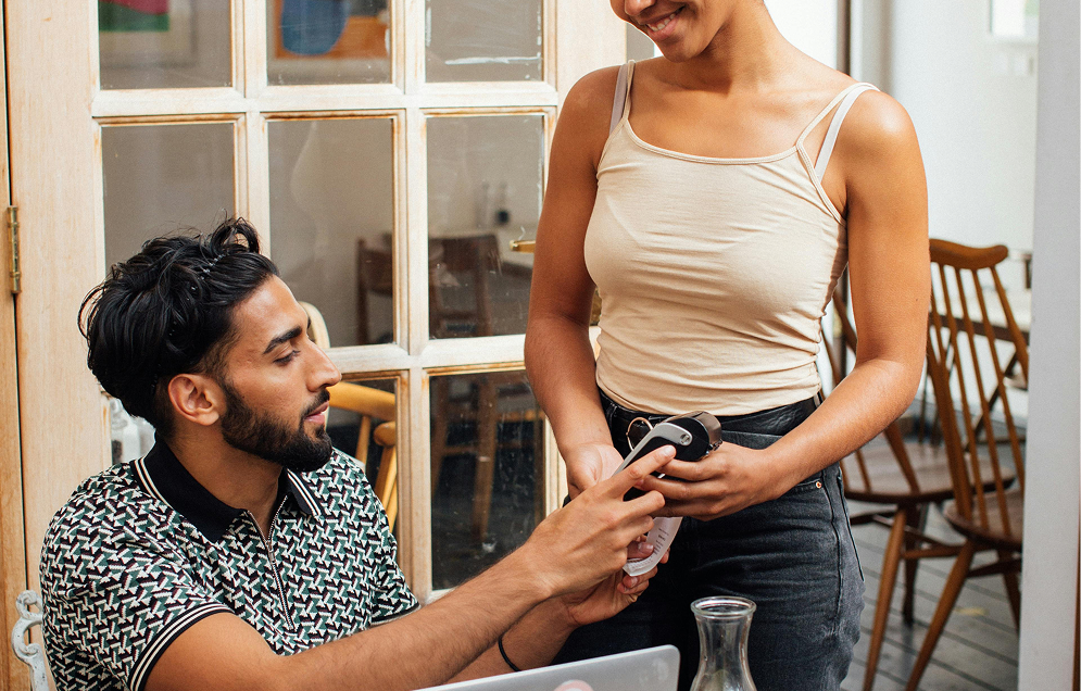 A smiling male baker in white uniform serving customers at a bakery counter with pastries on display and several people standing in line.