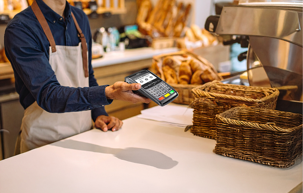 Modern restaurant interior with brick walls, gold pendant lights, wooden shelves stocked with goods, and diners seated at a table with a chef in the background.