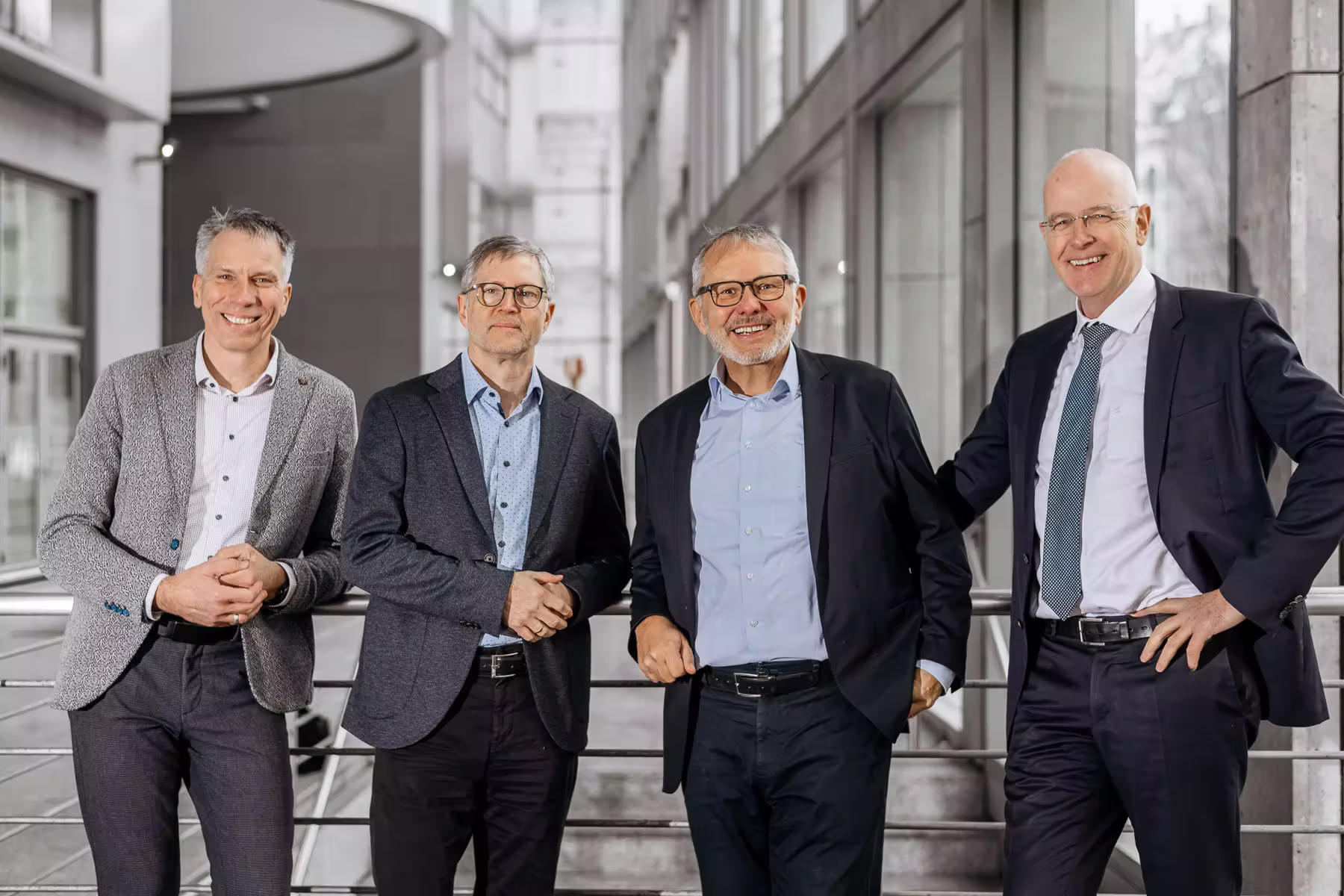Vier lächelnde Geschäftsfüherer in formeller Kleidung stehen nebeneinander in einem modernen Gebäude.