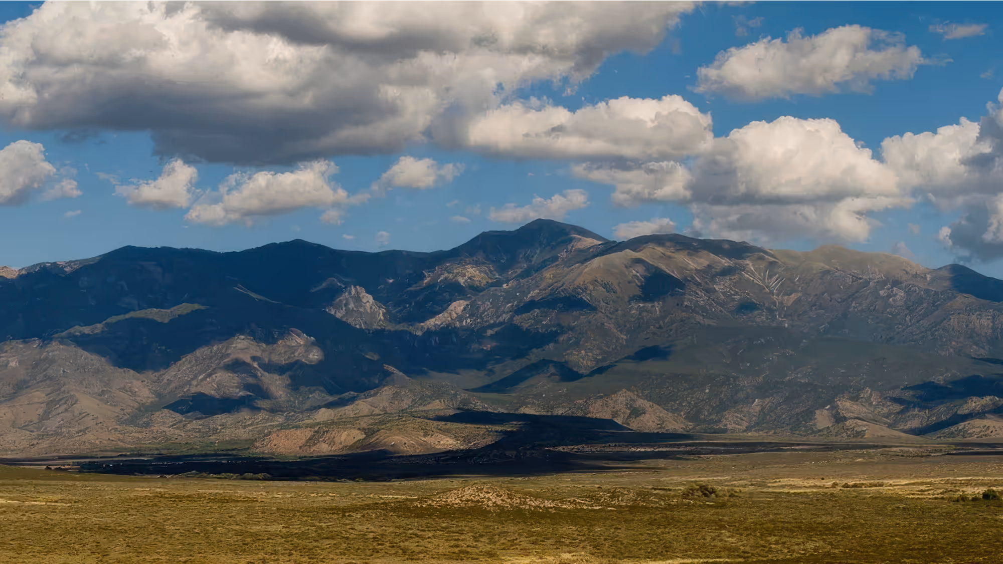 Expansive mountain range under a blue sky with scattered white clouds casting shadows on the terrain.