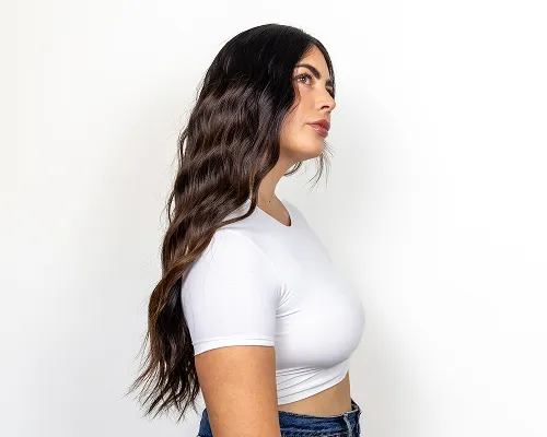 Ragazza di profilo con lunghi capelli castani mossi, top bianco e jeans su sfondo bianco