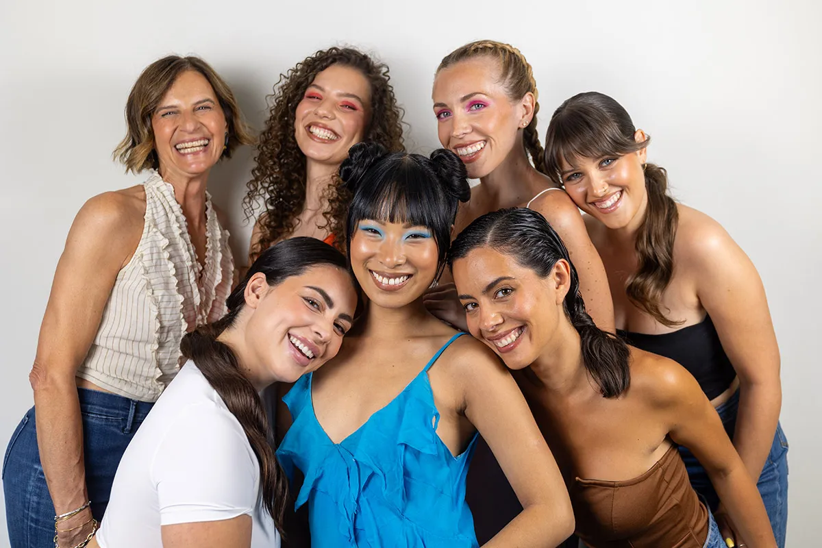 Gruppo di donne di età diverse sorridenti e abbracciate, con trucchi e acconciature colorate
