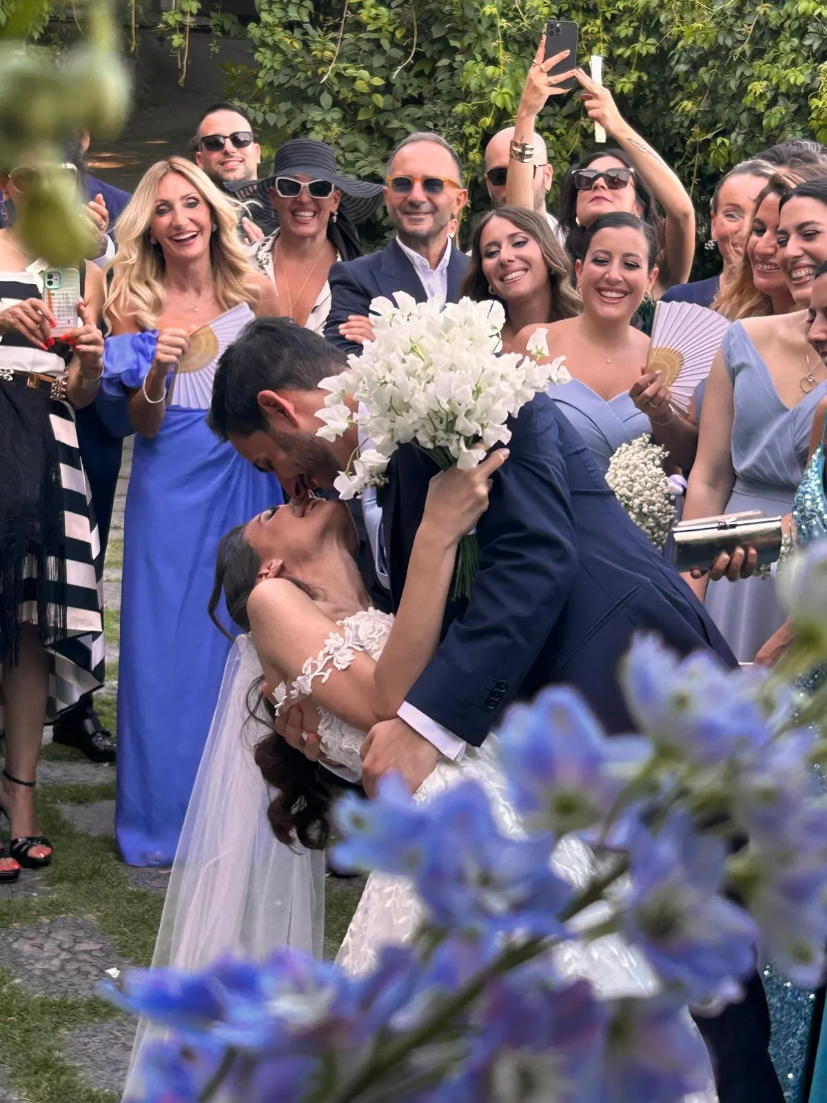 Sposi che si baciano circondati dagli invitati che li fotografano, con fiori azzurri in primo piano