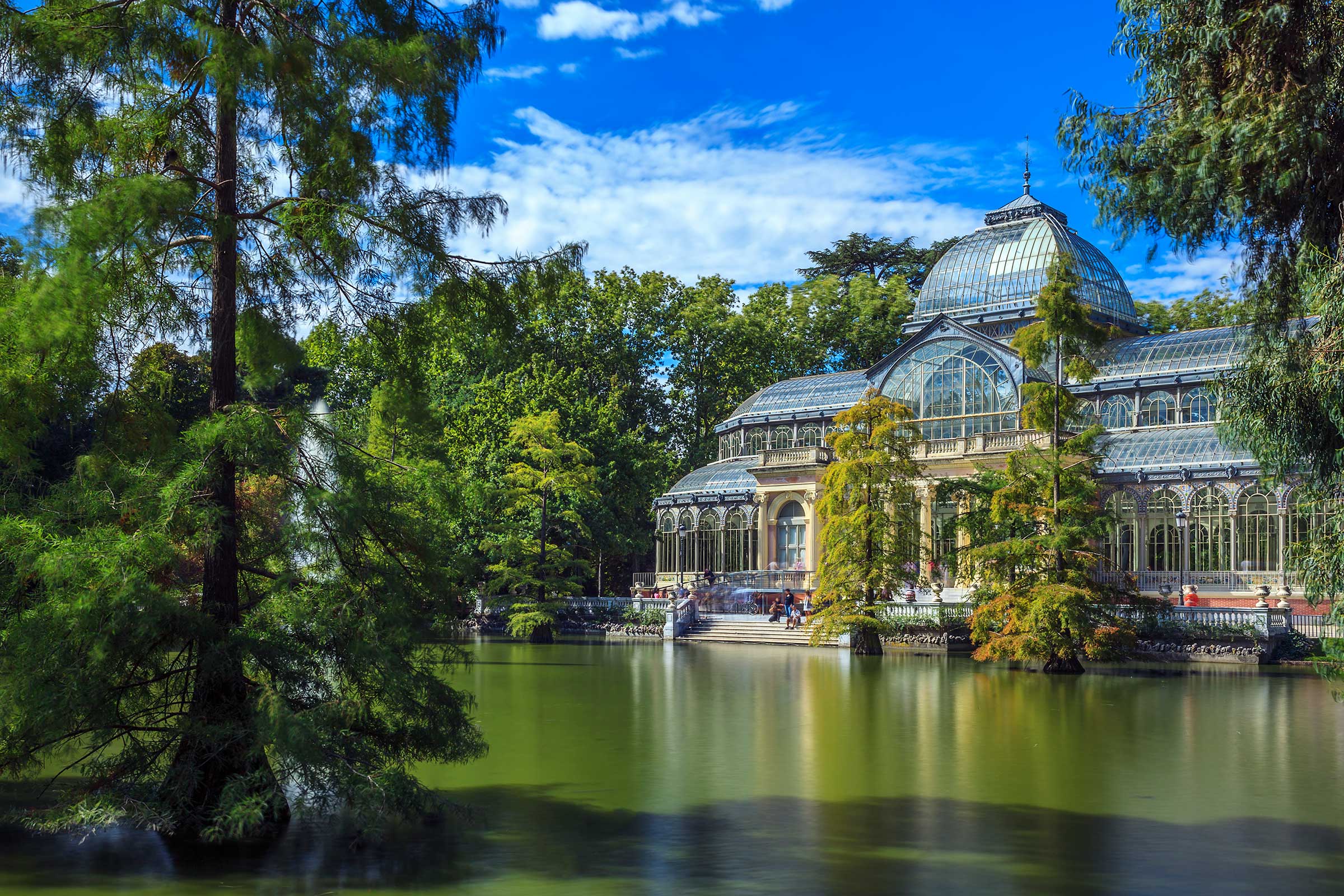 Glass conservatory building next to a green pond with trees and a blue sky overhead.