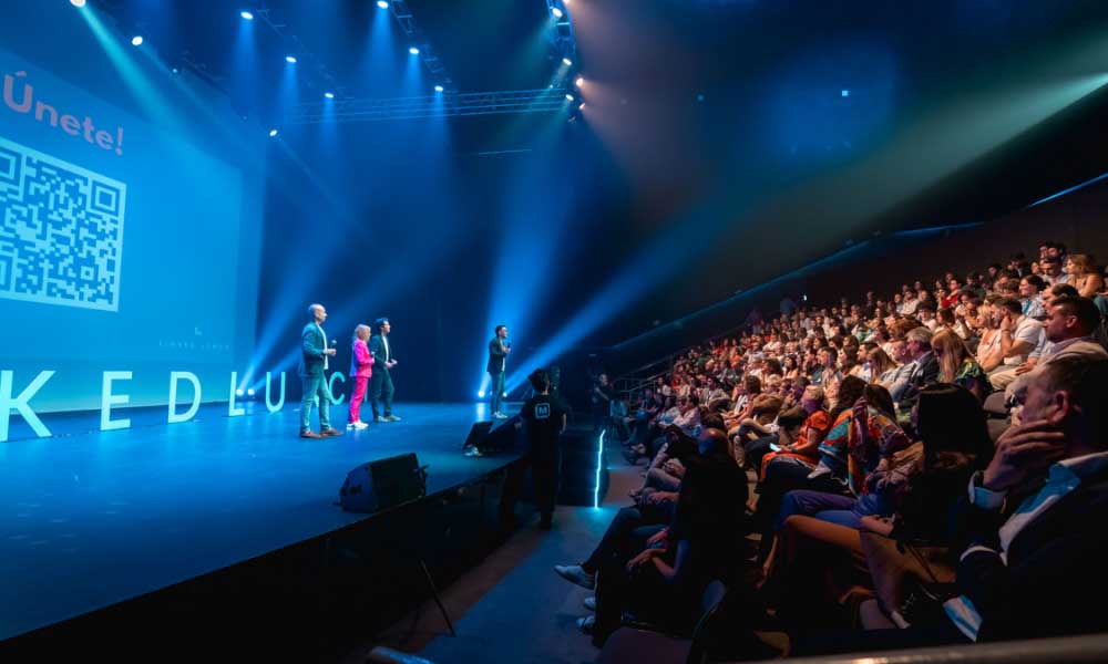 Four presenters on stage speaking to a large audience seated in a dimly lit auditorium with blue lighting and a QR code displayed on a screen.