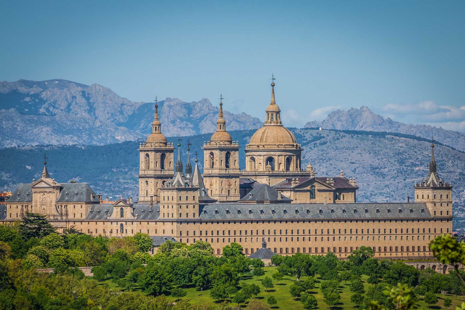 Large historic monastery with multiple towers and a central dome set against a mountainous background and surrounded by green trees.