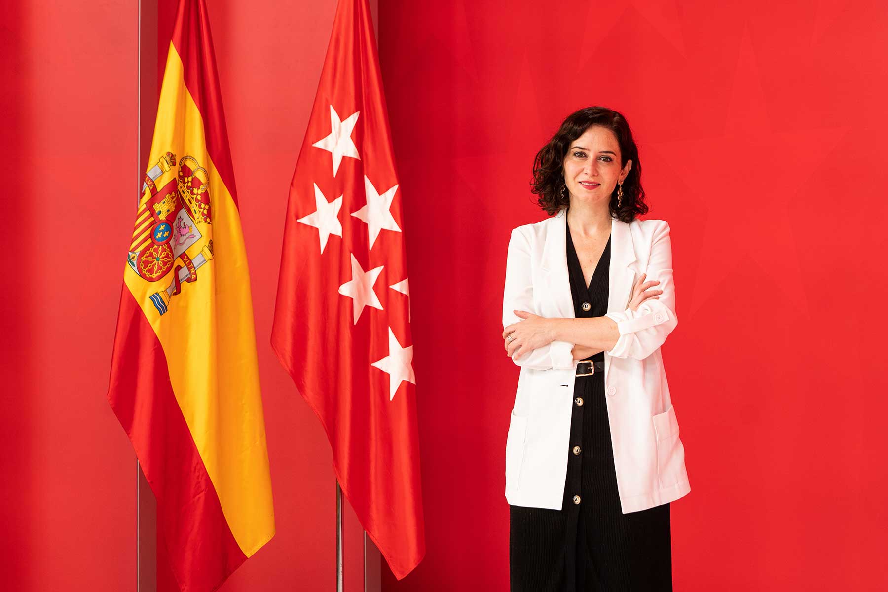Woman with dark hair wearing a white blazer over a black dress stands with arms crossed next to Spanish and regional flags against a red wall.