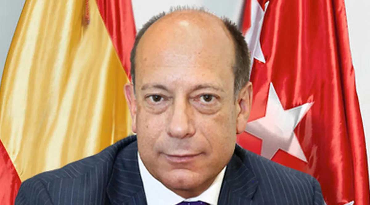 Middle-aged man in a striped suit and purple tie with Spanish and Madrid flags in the background.