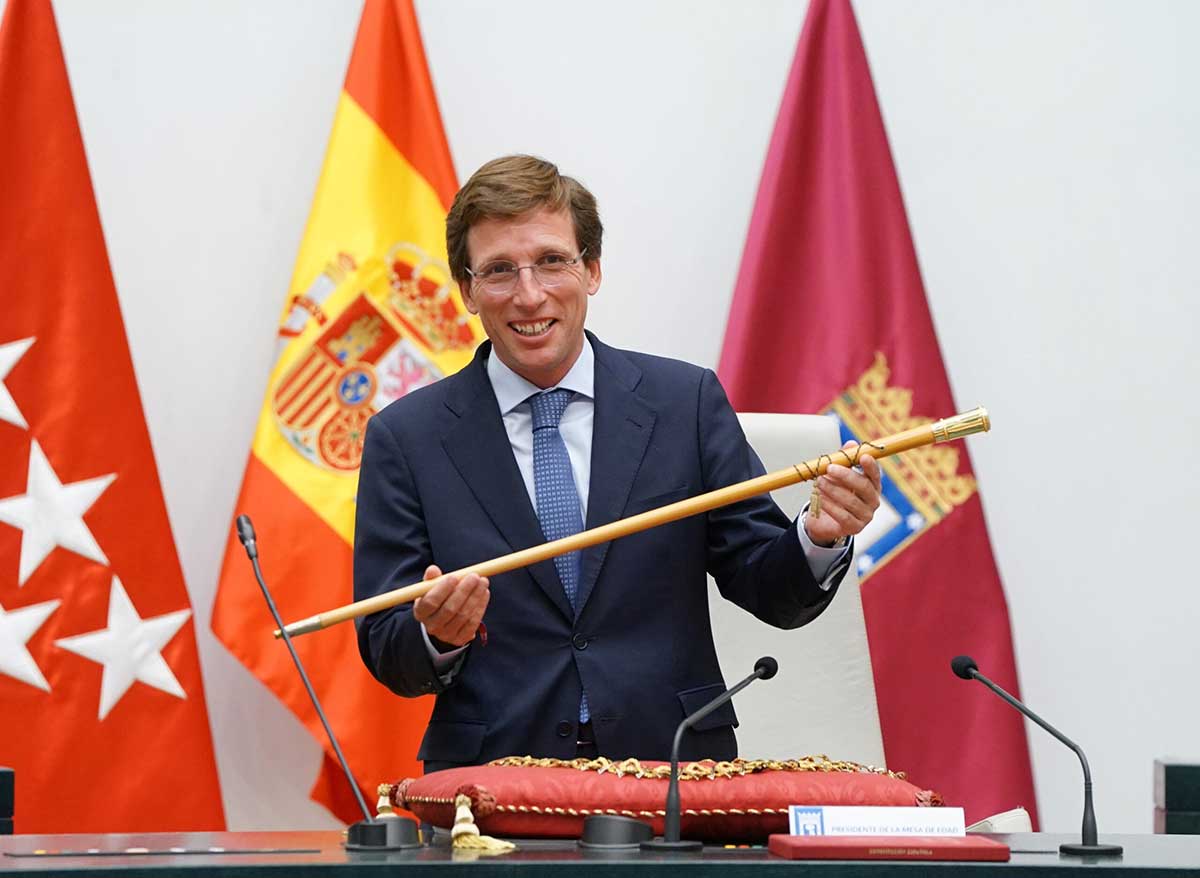 Man in suit and tie holding a ceremonial wooden staff, standing behind a table with microphones and red cushion, with Spanish and regional flags in the background.